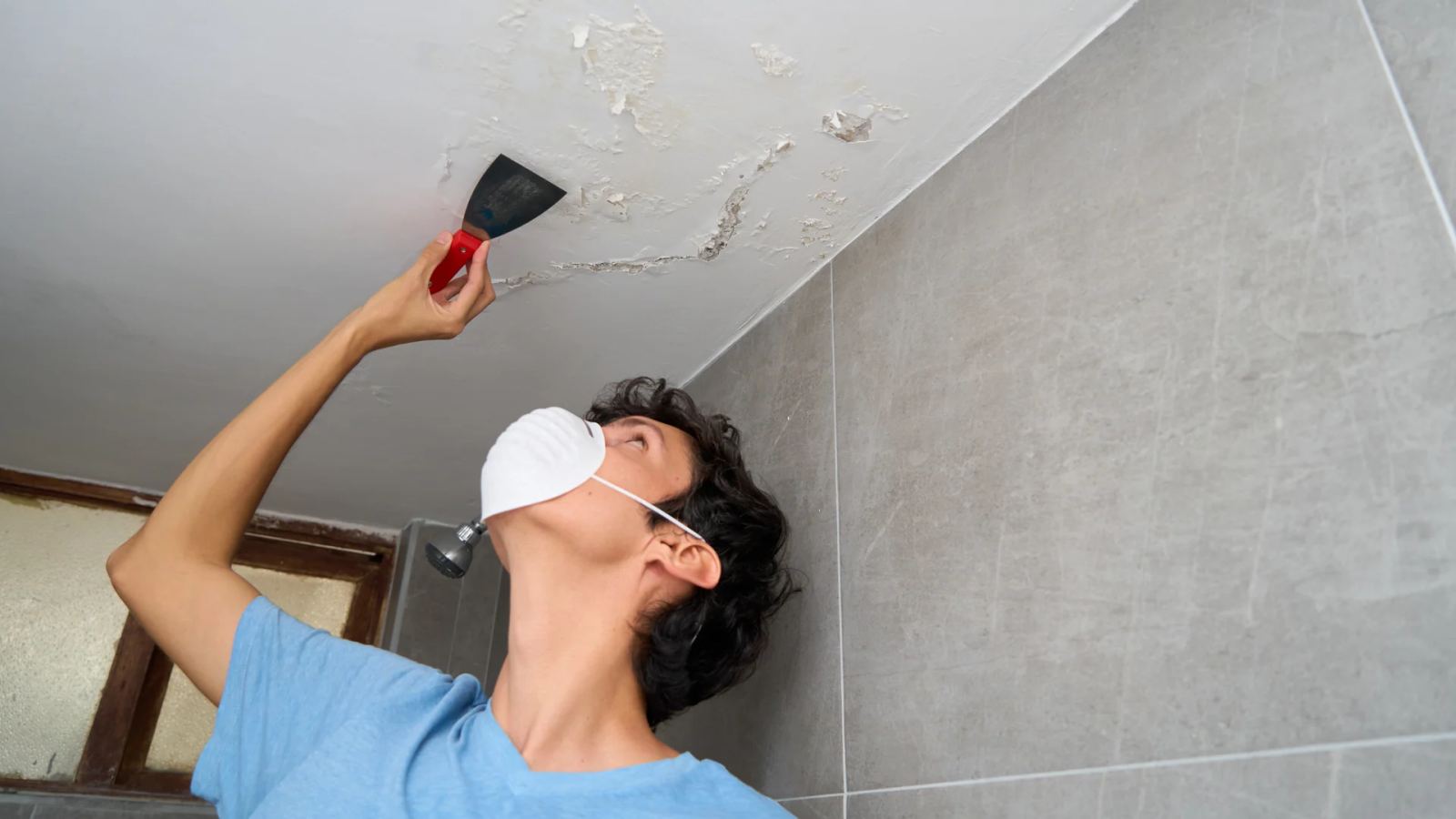 Person in a dust mask scraping water-damaged paint from a bathroom ceiling